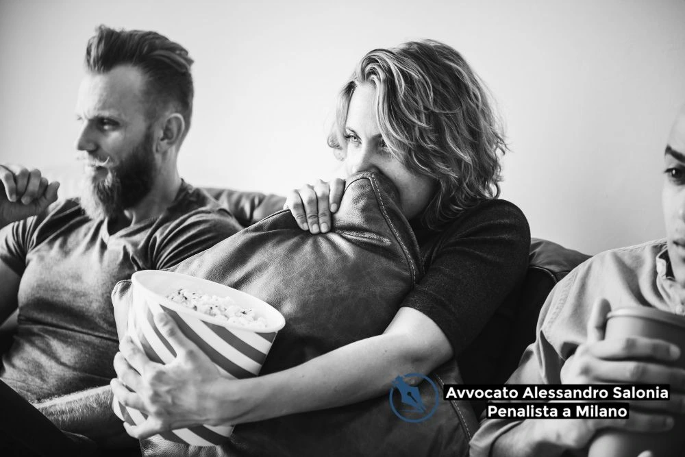 Tre persone sedute su un divano in un'immagine in bianco e nero: una donna al centro stringe un cuscino coprendosi parte del volto e mostra uno sguardo teso o preoccupato. In basso a destra compare il logo dello Studio Legale Alessandro Salonia, Avvocato Penalista a Milano.